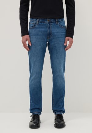 Mann trägt gerade geschnittene blaue Jeans, schwarzes langärmeliges Hemd und schwarze Lederschuhe, steht vor einem schlichten hellen Hintergrund.
