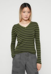 Young woman with straight ombre hair wearing an olive green and black striped V-neck sweater and gray cargo pants, hands in pockets.