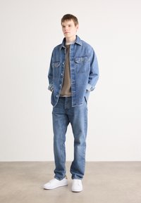 Jeansjacke mit zwei Brusttaschen, hellblau, mit Knopfverschluss, über einem beigen Pullover getragen, kombiniert mit passenden blauen Jeans und weißen Turnschuhen.