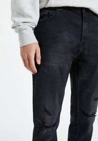 Czarne dżinsy z przetarciami, z dziurami przy kolanach i delikatną teksturą. Noszone z jasnoszarym swetrem.