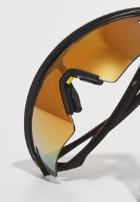 Sportowe okulary przeciwsłoneczne z zakrzywioną bursztynową soczewką, czarną oprawką, teksturowanym wykończeniem oraz elastycznymi ramionami z żółtymi akcentami i gumowymi uchwytami.