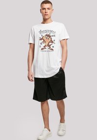 White cotton T-shirt with a graphic of the Tasmanian Devil. Worn with black athletic shorts and white sneakers. Simple design, casual fit.