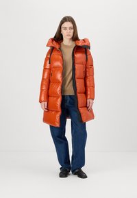 Puffer coat in bright orange with a shiny finish, featuring a hood, zip front, and black accents. Paired with a tan sweater and jeans.