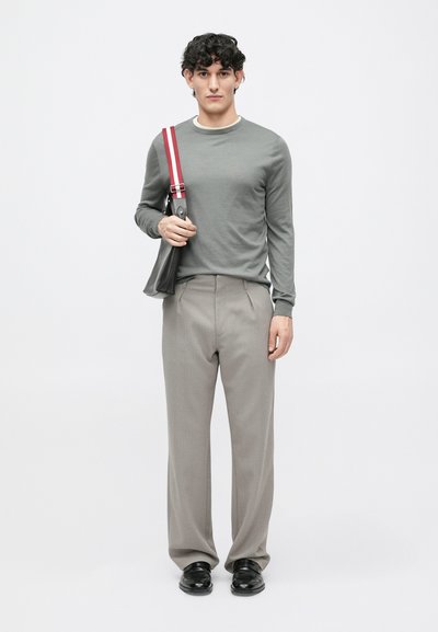 Graues Langarmhemd, hellbeige Rundhalsausschnitt, kombiniert mit hellgrauen Hosen. Schwarze Schuhe, eine schwarze Tasche mit roten Trageakzenten haltend.