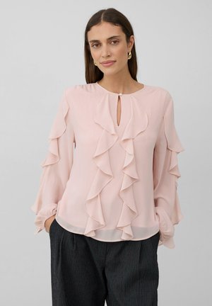 Frau in einer hellrosa, rüschengeschmückten Bluse mit langen Ärmeln und schwarzen karierten Hosen, die vor einem schlichten grauen Hintergrund steht.
