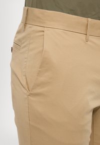 Beige kaki hlače z gladko teksturo, z ravnim sprednjim delom, stranskimi žepi in subtilnimi šivi. Lahke in prilegajoče se.