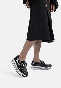 Scarpe con plateau in pelle verniciata nera, decorate con una catena argentata, dotate di una suola a righe bianche e nere e un design elegante con punta arrotondata.