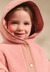 Różowy płaszcz z futerka syntetycznego o miękkiej fakturze, z dużym kapturem i kontrastową, gładką plisą z przodu. Wyposażony w okrągłe zapięcia na guziki.