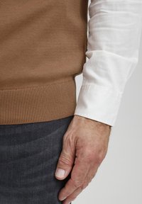 Brun stickad tröja med ribbad nederkant, parat med en vit skjorta. Handen vilar på mörkgrått tyg, vilket visar fingerdetaljer och textur.