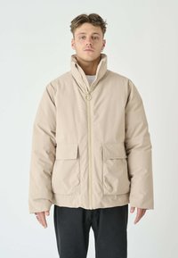 Helle beige, isolierte Jacke mit hohem Kragen, Frontrei�ßverschluss und zwei aufgesetzten Taschen. Glatte Textur, lockere Passform und minimales Design.