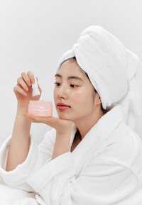 Barattolo rosa chiaro della maschera notte al collagene Abib PDRN tenuto con una presa delicata. Il modello indossa un accappatoio bianco e un asciugamano, mostrando un viso fresco.