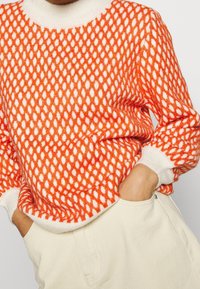 Pull en tricot avec un motif en losanges orange vif et blanc, doté d'un col rond et de poignets côtelés blancs. Texture douce.