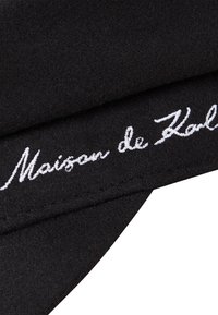 Chapeau en laine noir avec une texture douce, arborant un texte brodé blanc "Maison de Karl" sur le devant. Design classique avec une forme incurvée.