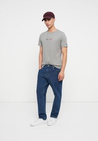 Γκρι βαμβακερό t-shirt με λογότυπο, συνδυασμένο με μπλε τζιν παντελόνι και λευκά αθλητικά παπούτσια. Το μοντέλο φοράει ένα μπορντό καπέλο. Απλή, casual εμφάνιση.