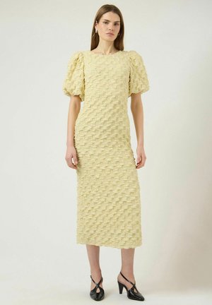 Kvinde iført en tekstureret, lysegul midi-kjole med pufærmer og sorte sko med hæl, stående foran en ensfarvet, lys baggrund.