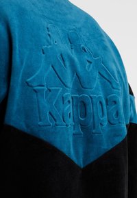 Blå och svart texturerad sweatshirt med en sammetsliknande finish, med en präglad "Kappa"-logotyp och grafik på baksidan.