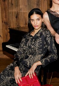 Zwarte satijnen top met gouden bloemenprint, lange mouwen en knoopsluiting. Rode cadeau doos in de handen. Achtergrond toont een piano en een houten wand.