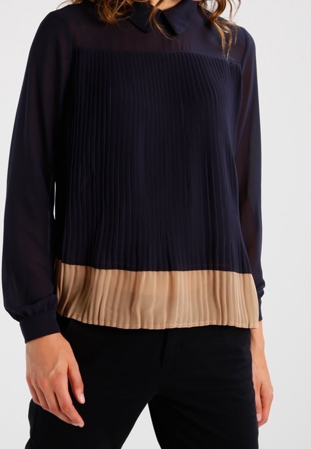 Femme portant un chemisier noir à manches longues avec un ourlet beige plissé et un col noir, associé à un pantalon noir.