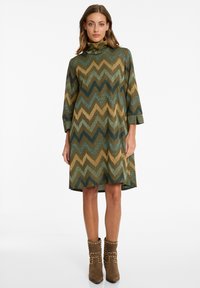 Robe midi avec un col montant, présentant un motif en zigzag dans des tons de vert, or et marron. Fabriquée en tissu texturé et brillant.
