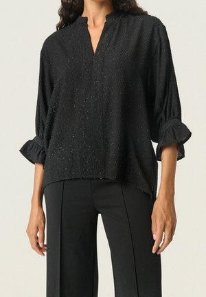 Frau trägt eine schwarze, strukturierte Bluse mit dreiviertellangen Rüschenärmeln und schwarze, maßgeschneiderte Hosen vor neutralem Hintergrund.