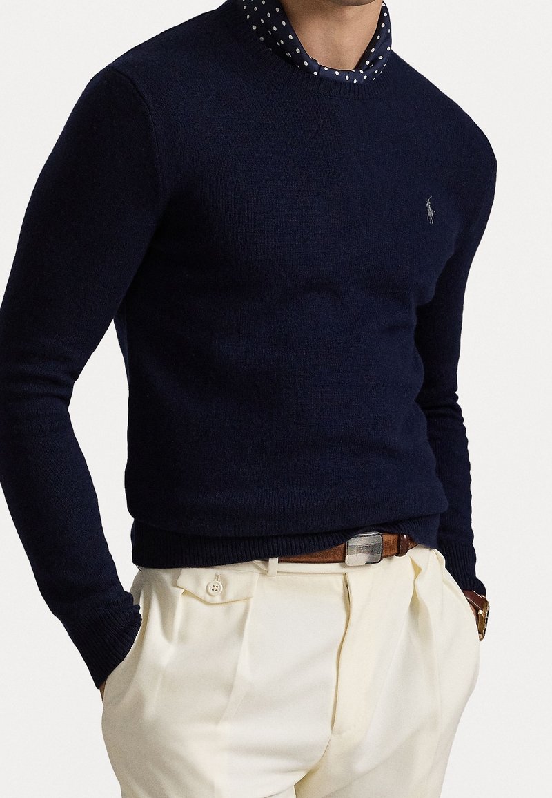 Homme portant un pull bleu marine par-dessus une chemise bleu marine à pois, un pantalon beige plissé, une ceinture marron et une montre-bracelet, les mains dans les poches.