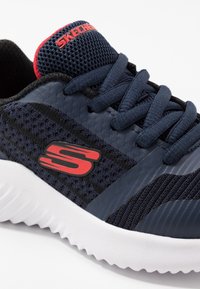 Baskets de sport marines et noires avec une tige en mesh, accents rouges et semelle extérieure blanche. Présente des lacets plats et un logo sur le côté.