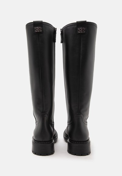 Paire de bottes hautes en cuir noir avec talons épais, fermetures éclair visibles à l'arrière et petits logos métalliques décoratifs près des bords supérieurs.