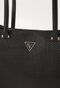 Geantă tote țesută neagră, cu bretele din piele, având o plăcuță logo triunghiulară în tonuri de aur. Suprafață texturată cu un finisaj elegant.