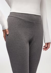 Leggings grigi in tessuto elastico, con vita alta e superficie liscia. La mano del modello è appoggiata sul lato del leggings.