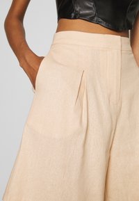 Pantaloni beige a vita alta realizzati in tessuto testurizzato, con pieghe frontali e tasche laterali. Il tessuto ha un aspetto morbido simile al lino.
