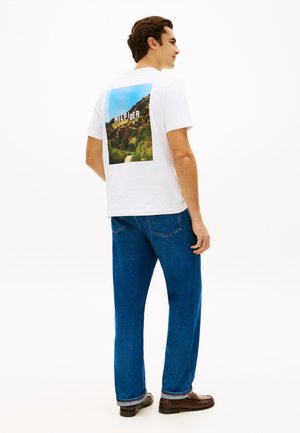 Άνδρας που φοράει λευκό T-shirt με το γράμμα "Hilfiger" στην πλάτη, μπλε τζιν και καφέ παπούτσια, στέκεται δεξιά σε λευκό φόντο.