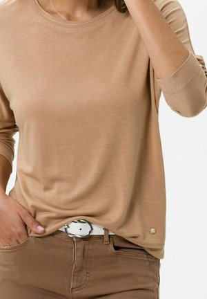 Beige Langarmshirt mit rundem Ausschnitt, weicher Textur und subtilen goldenen Akzent am Saum. Kombiniert mit hellbraunen Hosen und einem weißen Gürtel.