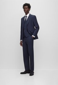 Marinefarbener Dreiteiler mit schmalem Revers, schmal geschnittenen Hosen und Knopfakzenten. Weißes Hemd und passender Krawatte, kombiniert mit dunklen Schuhen.