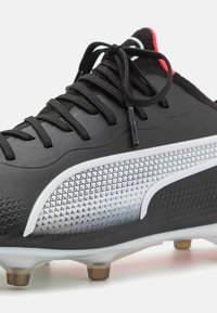 Botas de fútbol negras con parte superior texturizada, diseño degradado en blanco y detalles en rojo. Presenta cordones negros y tacos metálicos cónicos en la suela.