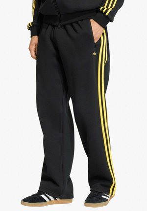 Pantaloni da tuta neri con strisce laterali gialle e piccolo logo, indossati con sneakers nere con strisce bianche e suole color gomma.