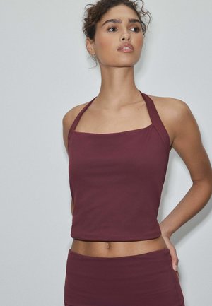 Žena v bezrukávovom bordovom halter crop tope a zodpovedajúcej sukni, mierne sa pozerajúca do strany na jednoduchom svetlom pozadí.