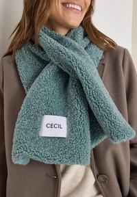 En teal fleece tørklæde med en tekstureret, fluffy design og et hvidt mærke med navnet "CECIL". Båret over en brun frakke.