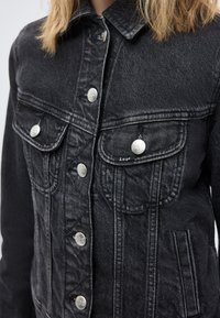 Μαύρο τζιν μπουφάν με ασημί κουμπιά με τη μάρκα Lee και τσέπες με καπάκι, φορεμένο από ένα άτομο με ξανθά μαλλιά μέχρι τους ώμους.