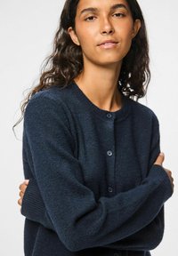 Tumši zils adīts kardigans ar apaļu kakla izgriezumu, garām piedurknēm un pogu aizdari. Iezīmējas ar mīkstu struktūru un ribotiem piedurkņu galiem.