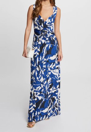 Kobieta w niebiesko-biało-czarnej kwiecistej maxi sukience bez rękawów z dekoltem w serek i paskiem w talii, trzymająca białą kopertówkę.