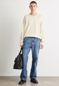 Κρεμ πλεκτό πουλόβερ με ριγωτές μανσέτες, συνδυασμένο με ανοιχτό μπλε τζιν παντελόνι. Το μοντέλο κρατά μια μαύρη δερμάτινη τσάντα, στέκεται σε μια ουδέτερη επιφάνεια.