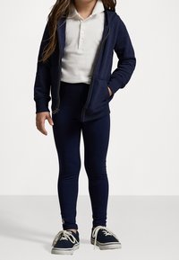 Dunkelblaues Zip-Hoodie über einem weißen Poloshirt, kombiniert mit figurbetonten, dunkelblauen Leggings und dunkelblauen Sneakers mit weißen Schnürsenkeln. Schlichtes Design, lässiger Stil.