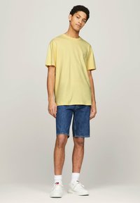 Camiseta amarilla de manga corta con un logo en el pecho, combinada con shorts de mezclilla azules y zapatillas blancas. El modelo adopta una expresión neutral.