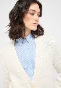 Krämfärgad stickad cardigan med V-ringning, öppen front och detalj med en enda knapp, bärs över en ljusblå randig skjorta.