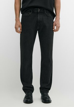 Mann trägt schwarze Jeans mit geradem Bein, schwarze Lederschuhe und ein schwarzes, in die Hose gestecktes Hemd und steht vor einem einfarbigen Hintergrund.