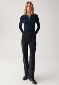Vrouw staande met handen achter de rug, gekleed in een marineblauwe ribgebreide lange mouwen henley, donkere hoog-taille broeken en zwarte schoenen.