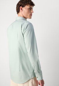 Cămașă lungă de culoare verde deschis, cu guler tip spread, manșete cu nasturi și detalii pe cusătura din spate; fabricată dintr-un material neted și ușor.