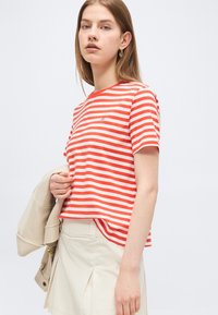 Young woman wearing red and white striped short-sleeve shirt and beige skirt, holding a beige jacket over one arm against a plain background.