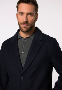 Cappotto di lana blu scuro con rever a punta, chiusura a un bottone e una polo grigia sotto. Linee pulite e texture liscia.