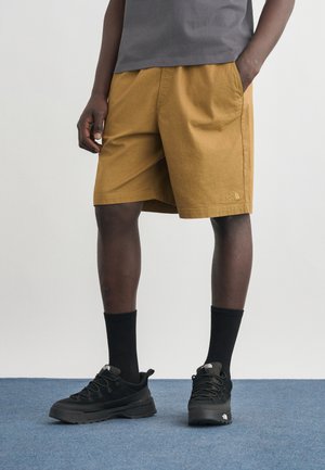 Mężczyzna w musztardowożółtych szortach, czarnych skarpetach crew i czarnych sportowych butach stojący na niebieskim dywanie, z rękami w kieszeniach.